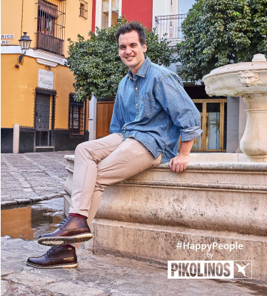 El periodista Julio Muñoz, sentado en una fuente en una plaza de Sevilla, sonriendo a la cámara y llevando un par de botines Pikolinos en color marrón.