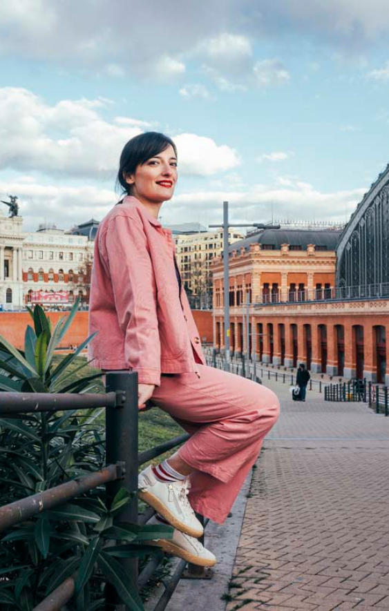Lara Lars, eine auf Collagetechnik spezialisierte Künstlerin, posiert auf einem Geländer durch die Straßen von Madrid. Sie trägt einen pinkfarbenen Anzug und weiße Pikolinos-Turnschuhe.