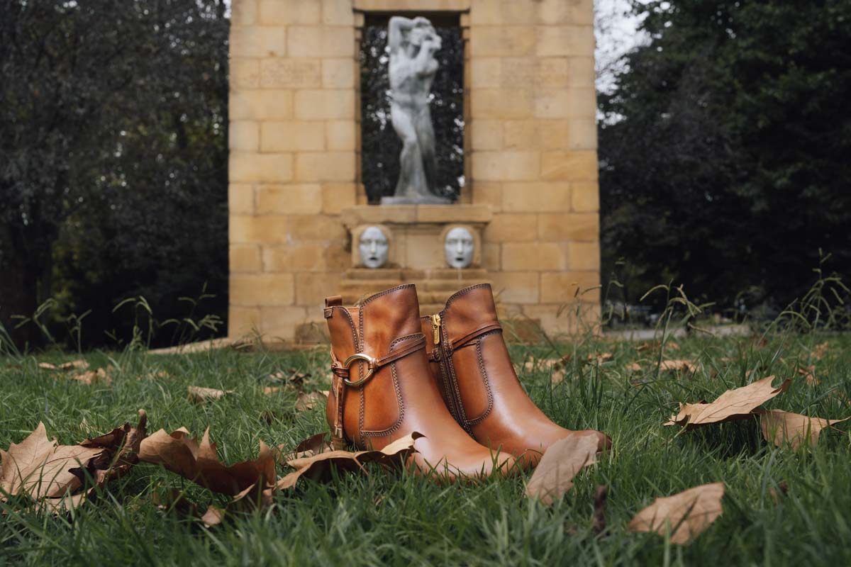 Photograph of a pair of brown Pikolinos women's ankle boots in the Doña Casilda park with a monument
                        in the background