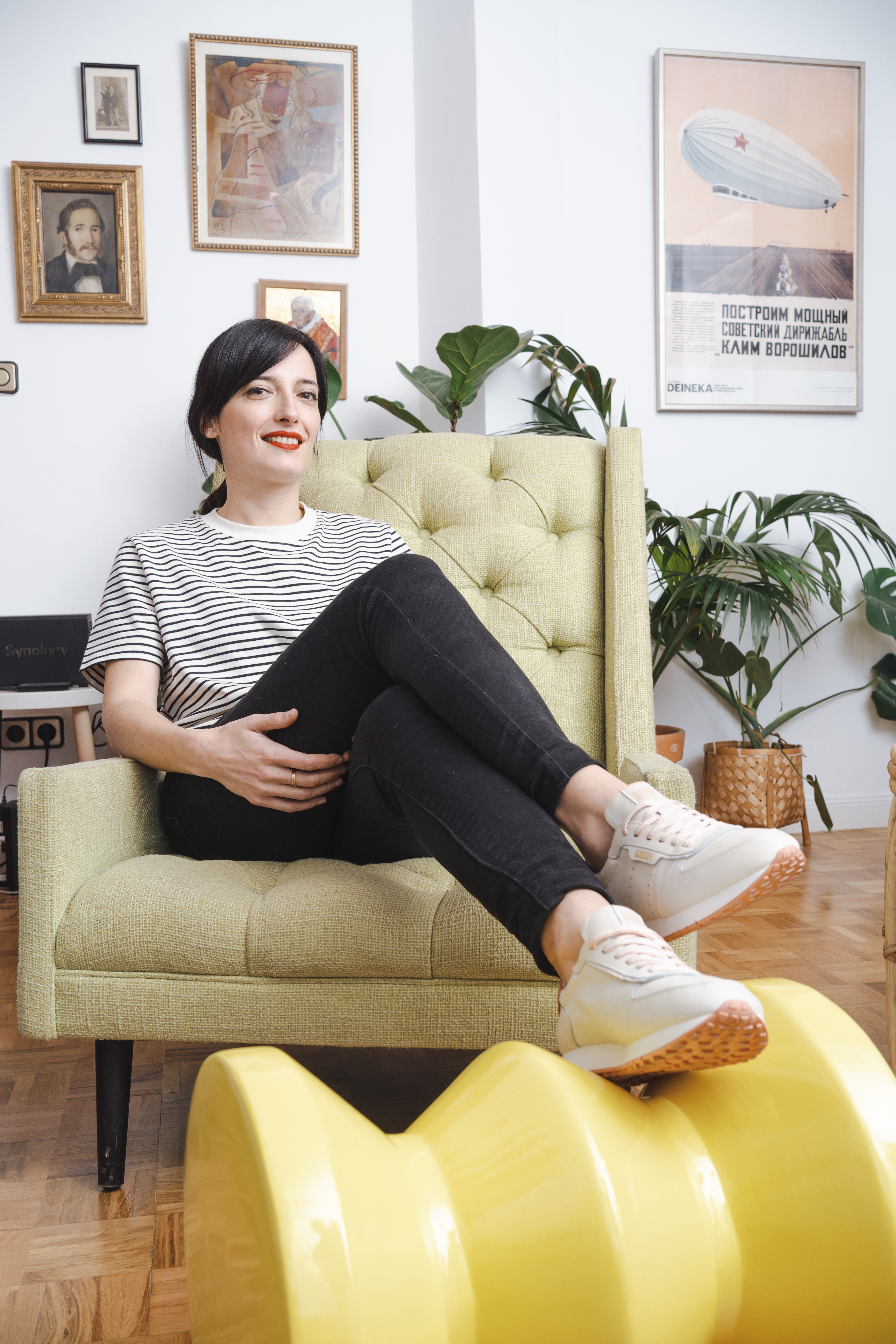 Lara Lars, sitting in an armchair with her legs crossed, supporting hers on a yellow sculpture. She shows her Pikolinos sneakers in white color and with an orange sole.