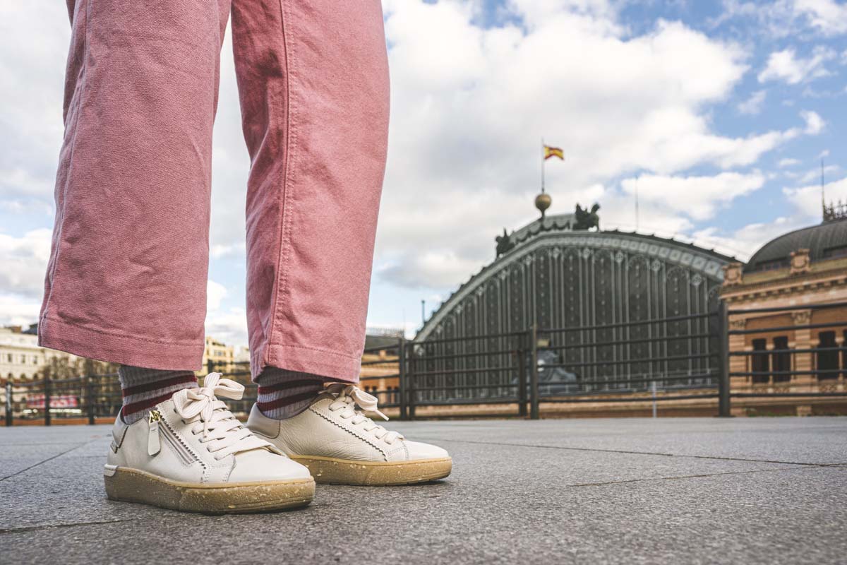 Image recadrée des pieds de Lara marchant dans la rue. Il porte un pantalon rose et des baskets blanches, avec des semelles couleur caramel.