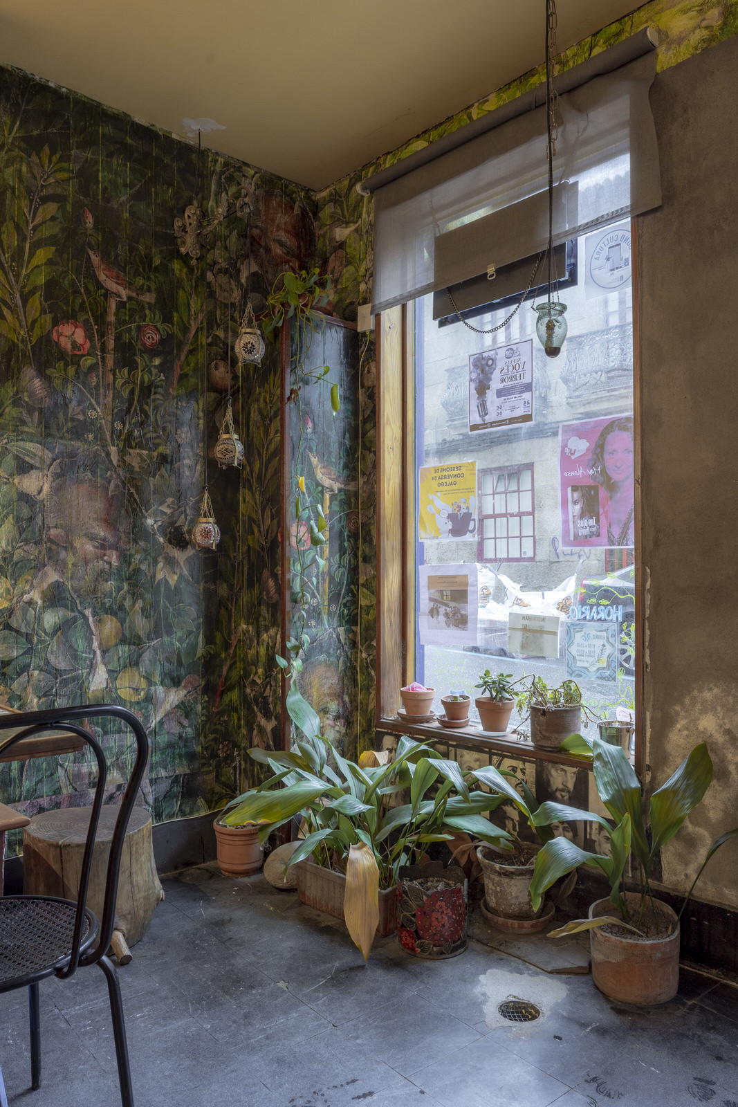 Imagen del interior de la cafetería, con un ventanal, plantas y una pared pintada de flores