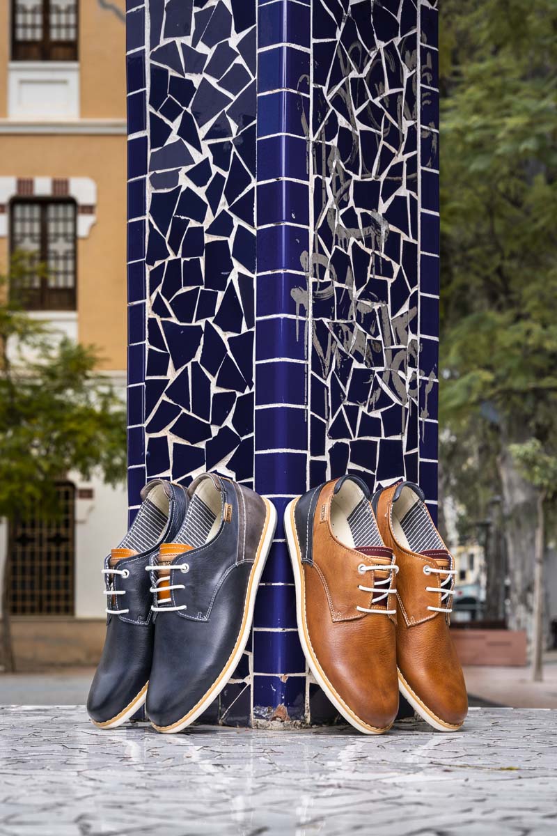 Picture of two models of men's dress shoes, one in blue and the other in brown, leaning against a column.