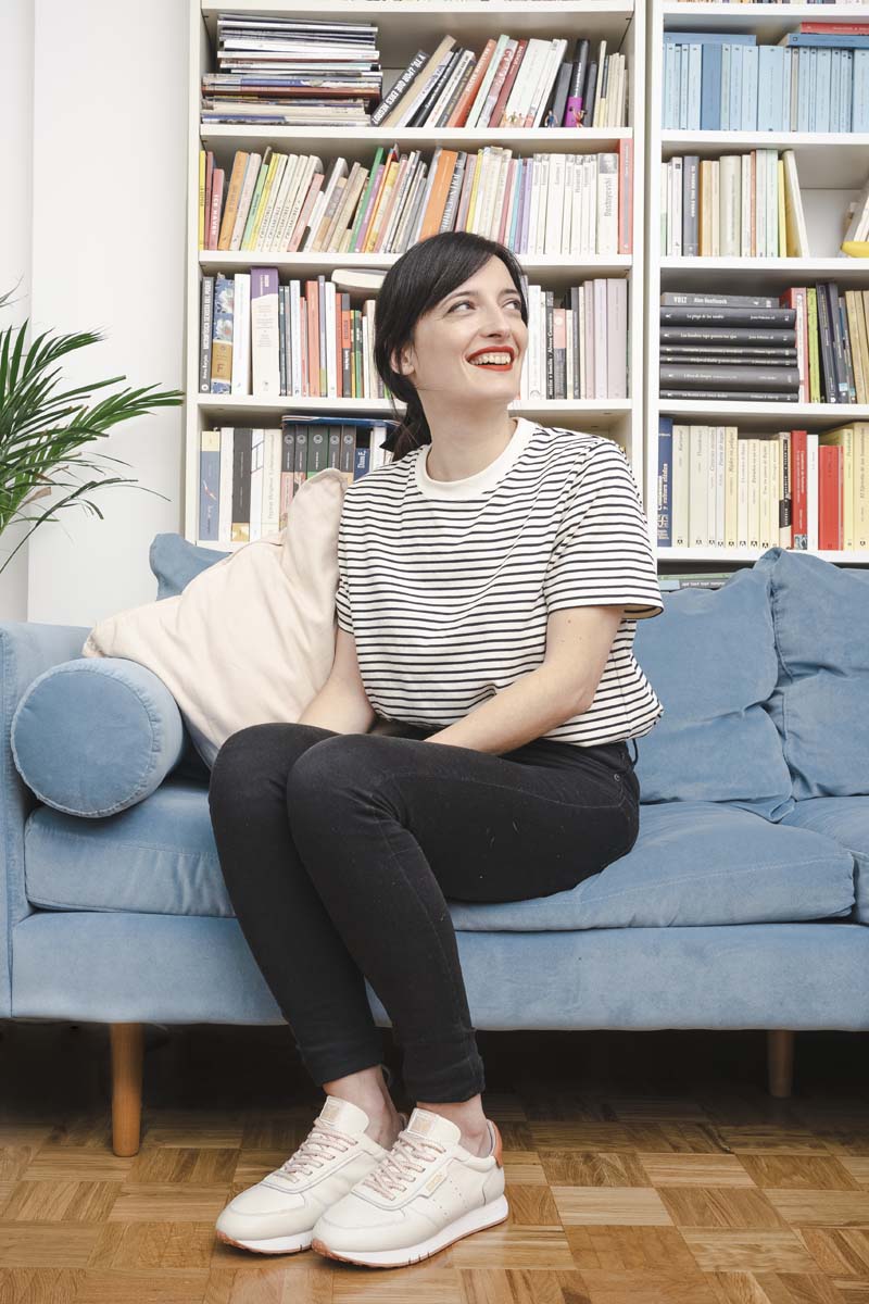 Lara Lars, assise sur le canapé à la maison, pose en souriant. Il porte une chemise rayée, un pantalon noir et des baskets Pikolinos blanches.