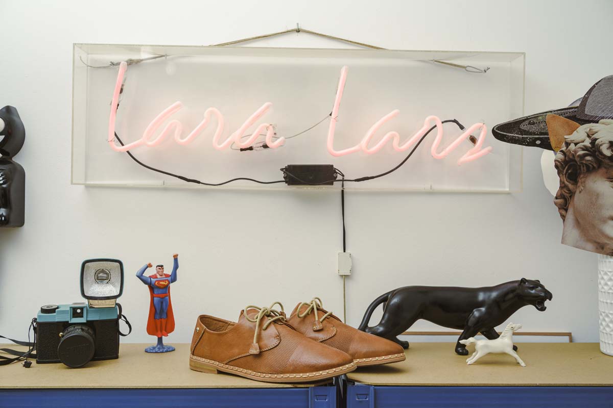 Ein Paar braune Pikolinos-Damenschuhe auf einem Möbelstück, umgeben von einer Fotokamera, ein paar Dekofiguren und einem Leuchtschild mit dem Namen Lara Lars.