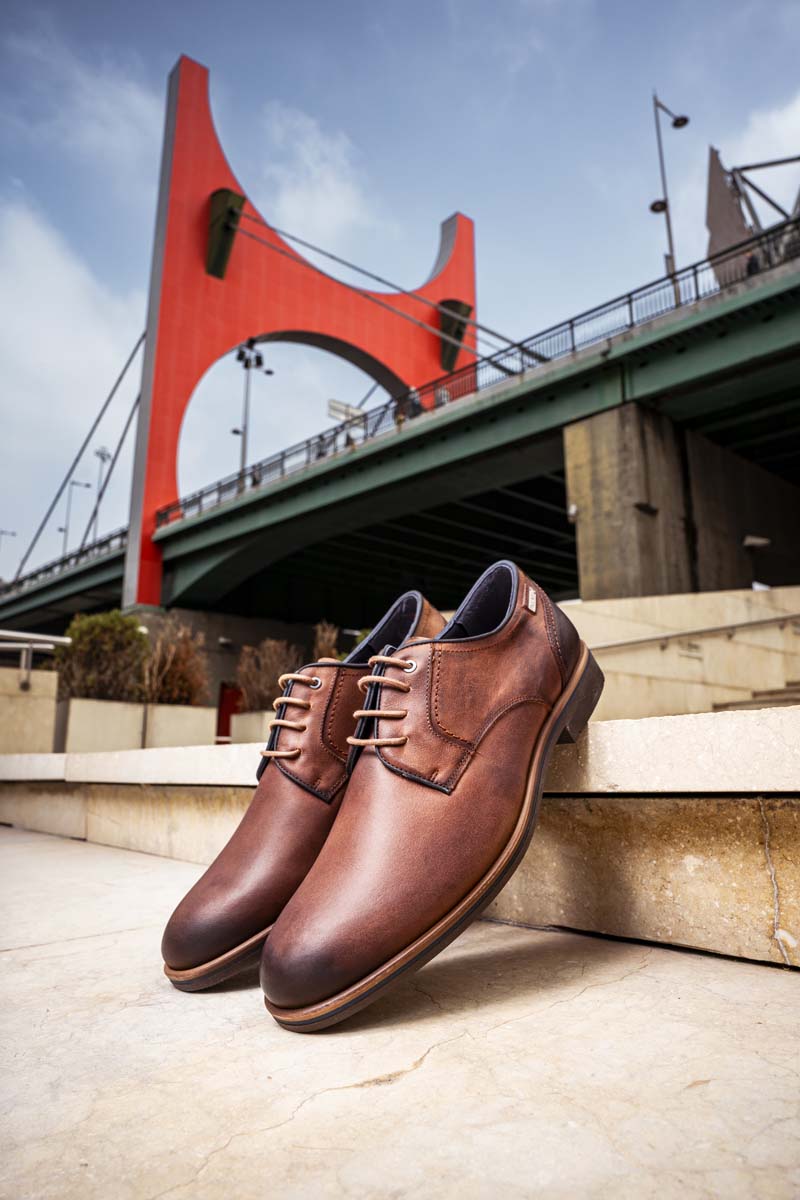 Fotografía de unos zapatos de vestir de Pikolinos de hombre color marrón en la calle con un puente de fondo 