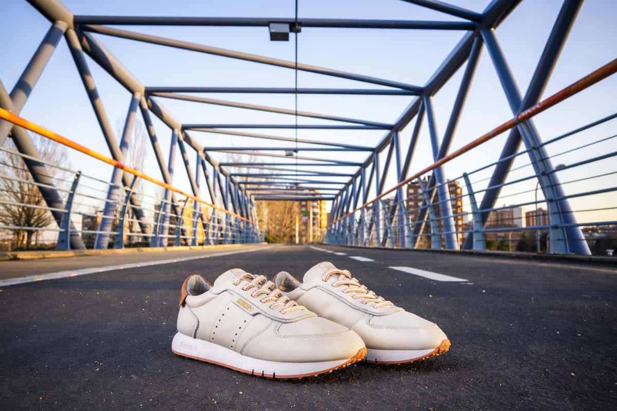 Still life image showing a pair of Pikolinos shoes. It is the Barcelona style in white color and it appears perched on the asphalt on a bridge in the city of Madrid.