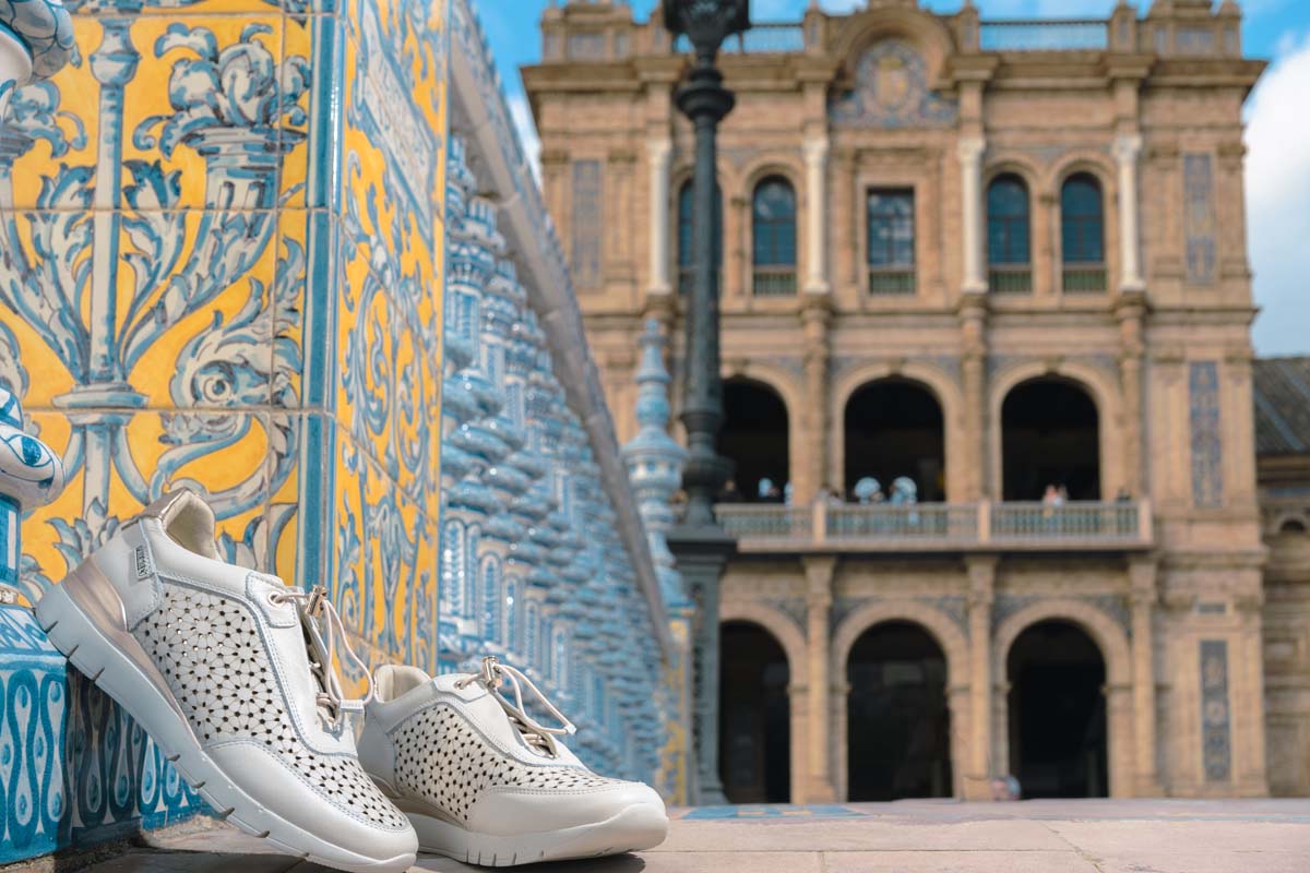 Photograph of women's sports shoes by Pikolinos leaning against a wall and the Plaza de
                        España in the background