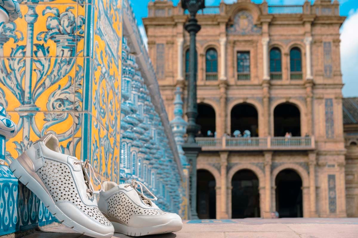 Photograph of women's sports shoes by Pikolinos leaning against a wall and the Plaza de
                        España in the background